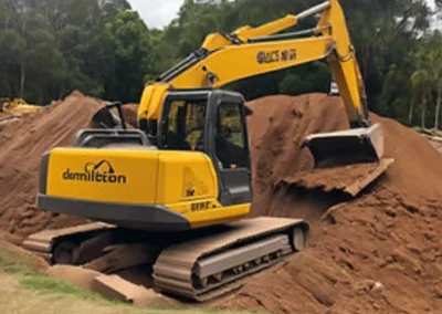 A yellow excavator moving dirt on a construction site with a large pile of earth and trees in the background. The word "demolition" is visible on the side of the excavator.